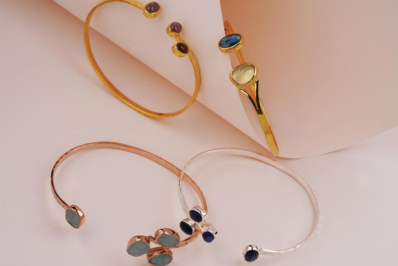 A close-up of 4 gold gemstone bangles, one white gold, two yellow gold, and one rose gold, on a pale peach backdrop.