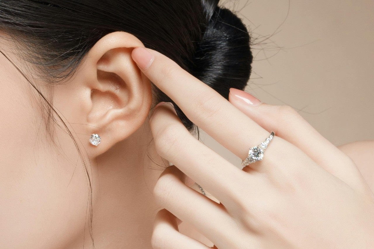A close-up of a young woman’s ear with a single diamond stud earring, emphasized by her finger, adorned with a glistening solitaire diamond ring.