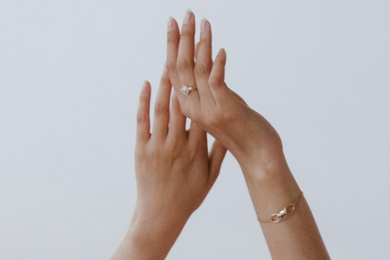 A close-up of a woman’s raised hands, a delicate chain bracelet on her right wrist and a diamond ring on her right middle finger.