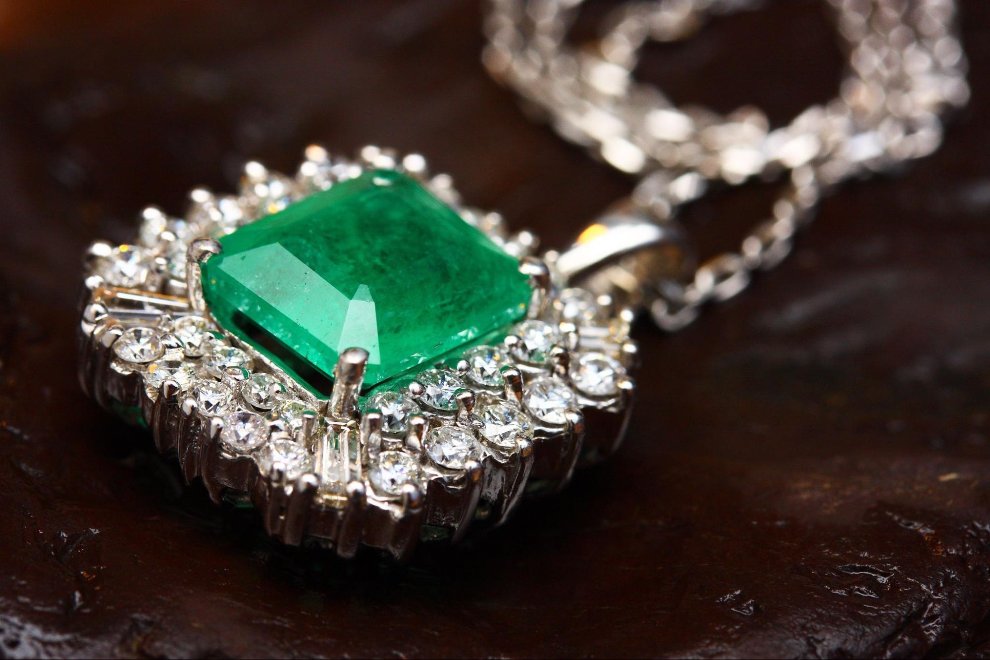 A close-up of a necklace pendant with a large green center stone and diamonds surrounding it laying on a dark wood surface.