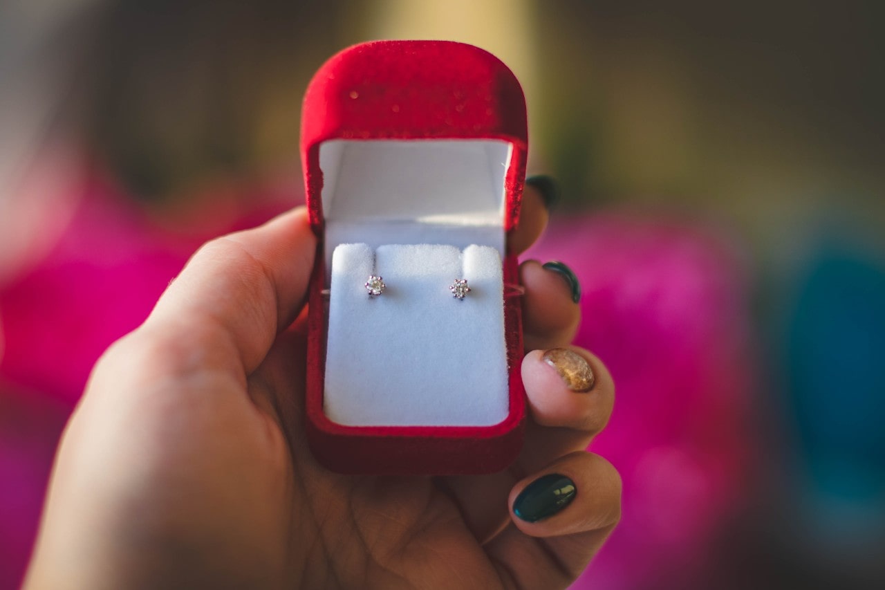 A close-up of a nicely manicured hand holding a red velvet earring box with diamond studs inside.