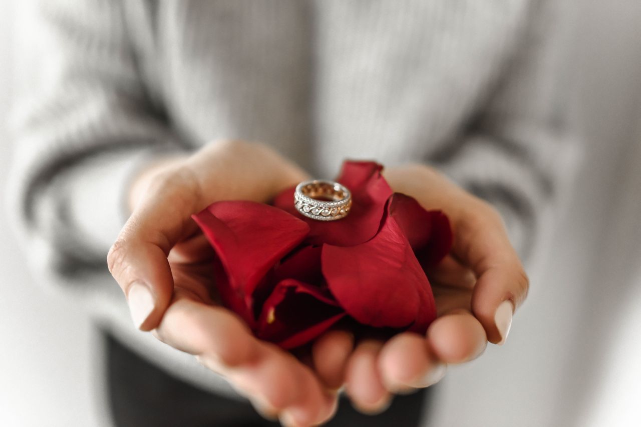 A close up of a person in a gray sweater holding a yellow gold wedding band with intricate detailing on top of red rose petals. A close up of a person in a gray sweater holding a yellow gold wedding band with intricate detailing on top of red rose petals.