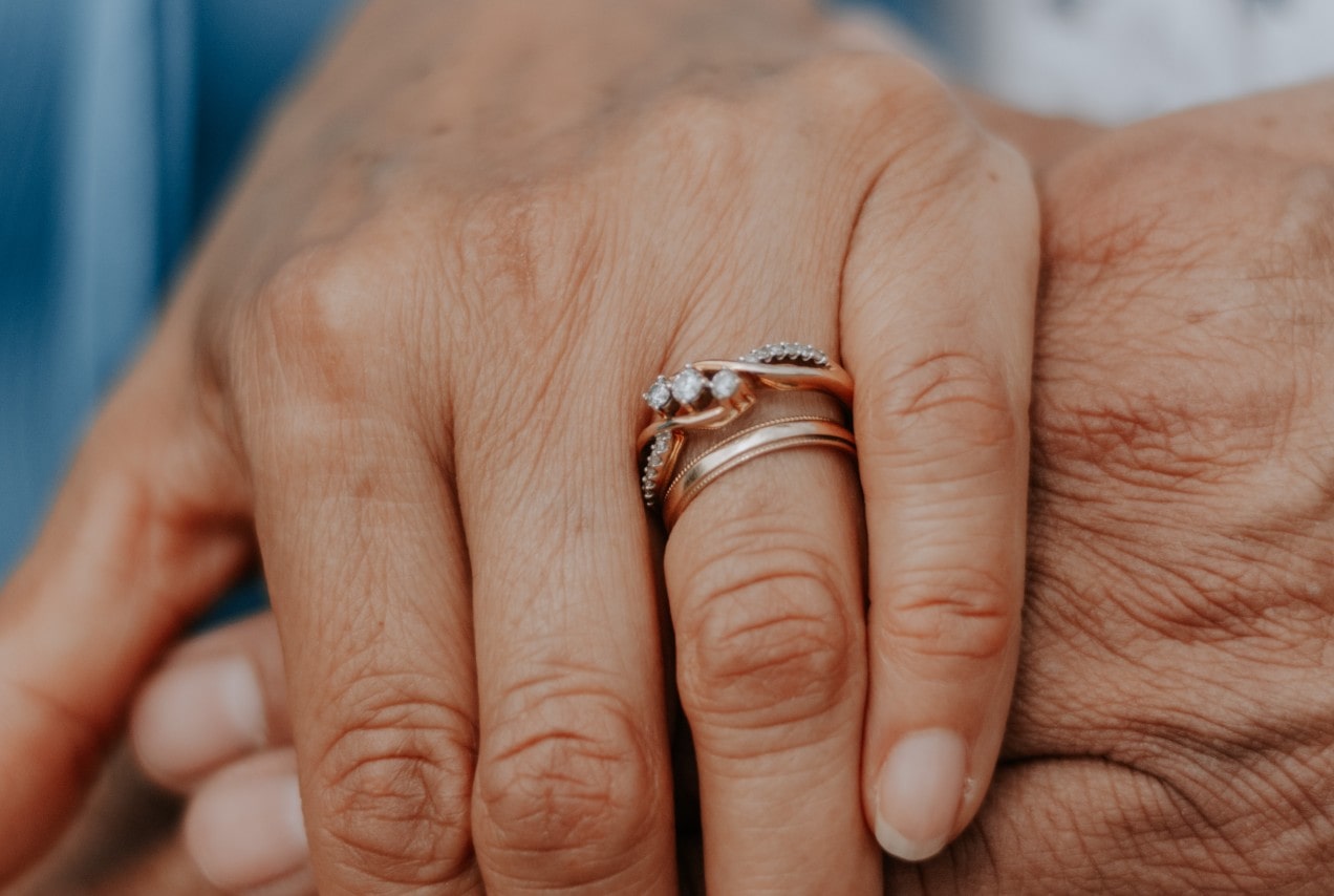 A close up of two aged hands holding each other, showcasing delicate nature-inspired rose gold wedding bands on a finger. A close up of two aged hands holding each other, showcasing delicate nature-inspired rose gold wedding bands on a finger.