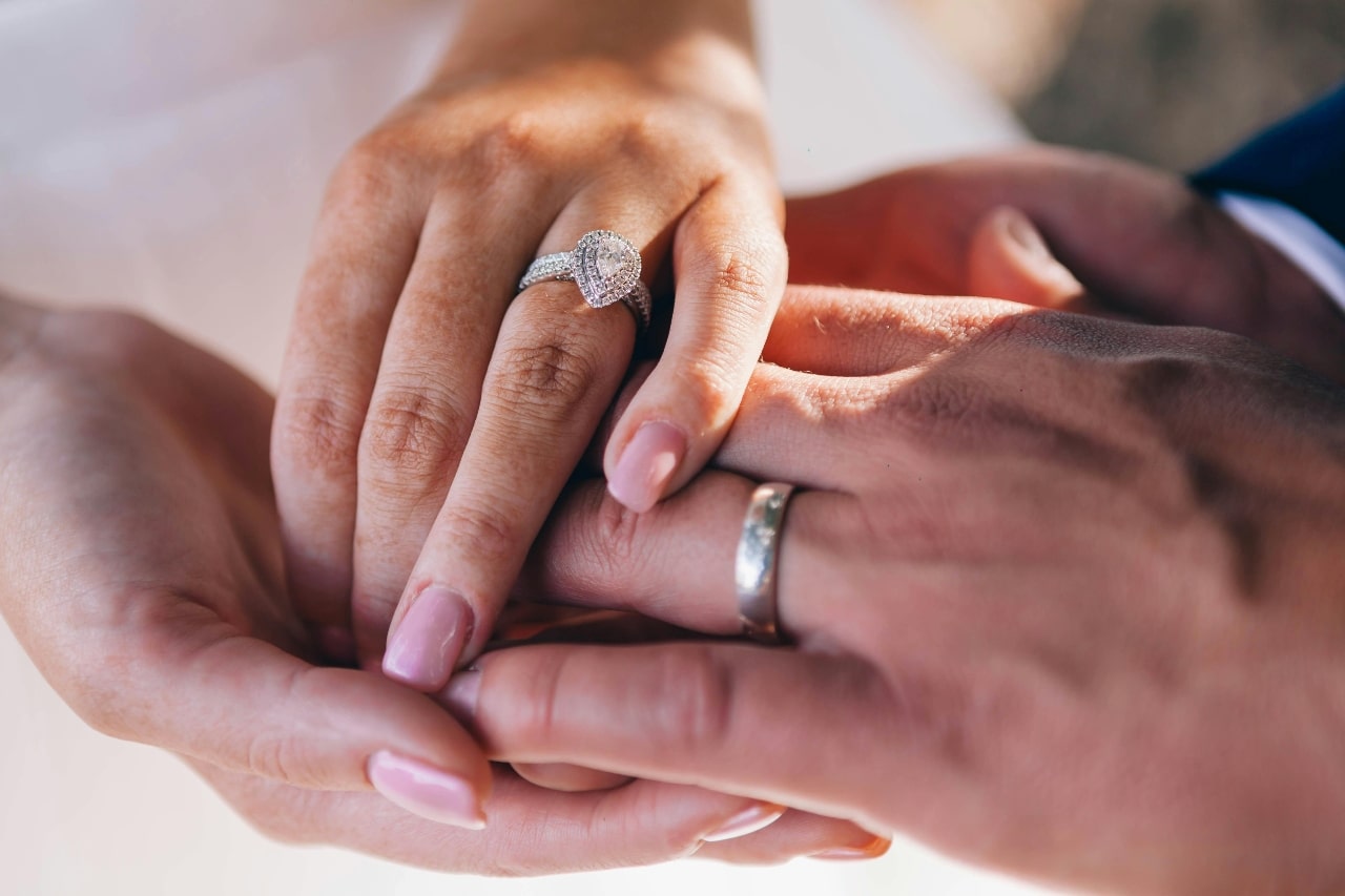 A close up of an intertwined hands, showcasing a white gold diamond pear shaped halo engagement ring