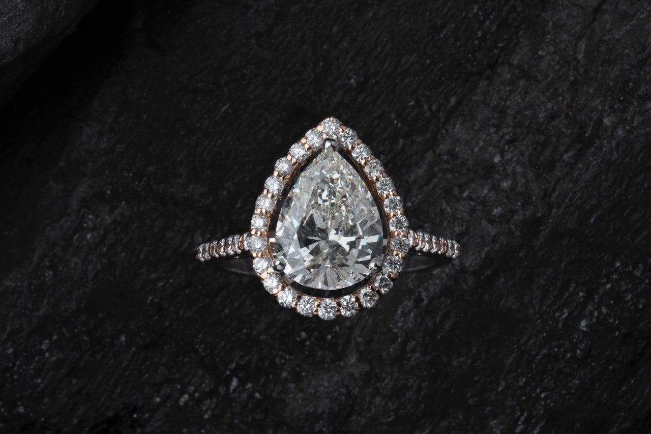 A pear-shaped diamond ring with a halo of smaller diamonds, set against a dark textured background.