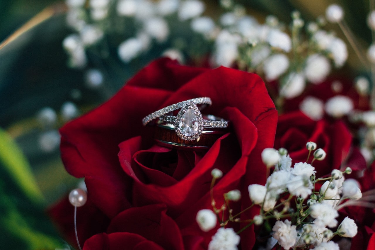 A pear-shaped diamond ring nestled in a vibrant red rose, surrounded by delicate white baby's breath flowers.