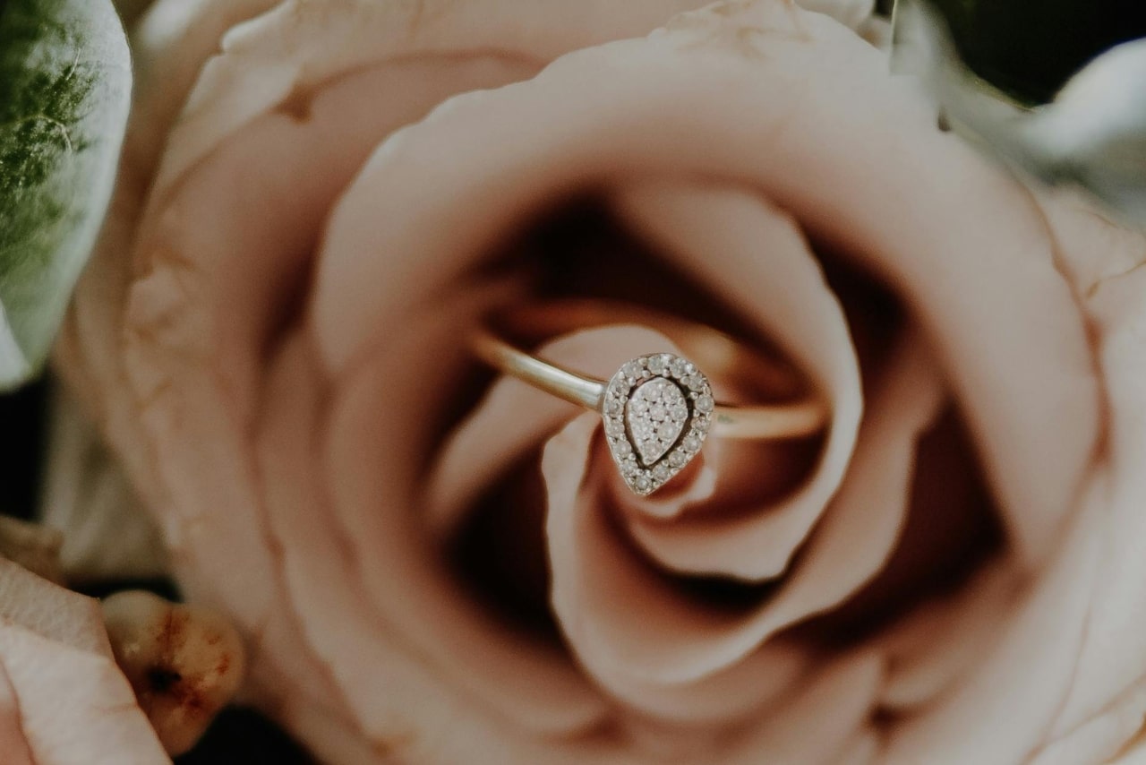 A pear-shaped diamond ring with a gold band is elegantly placed in the center of a soft pink rose.