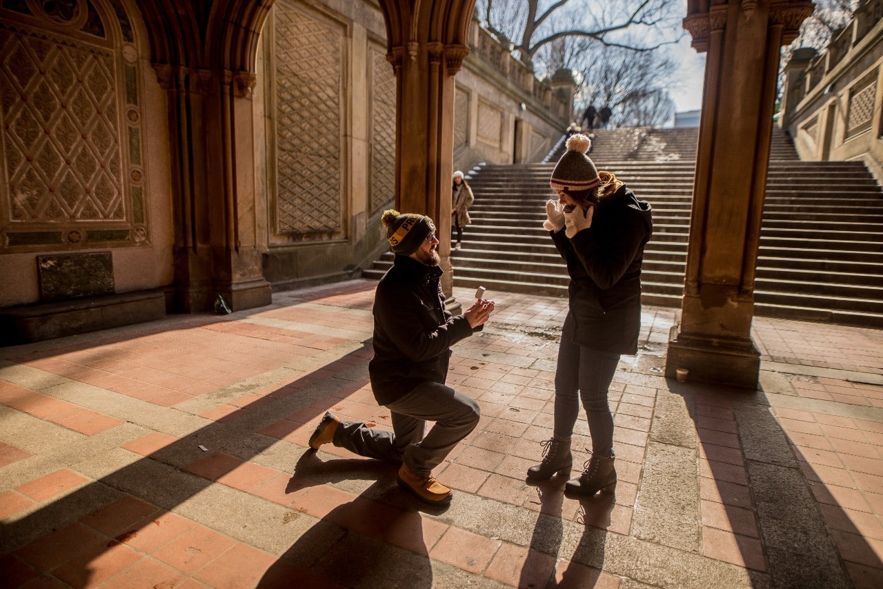 A man kneels to propose to a surprised woman under an ornate archway, with sunlight casting shadows.