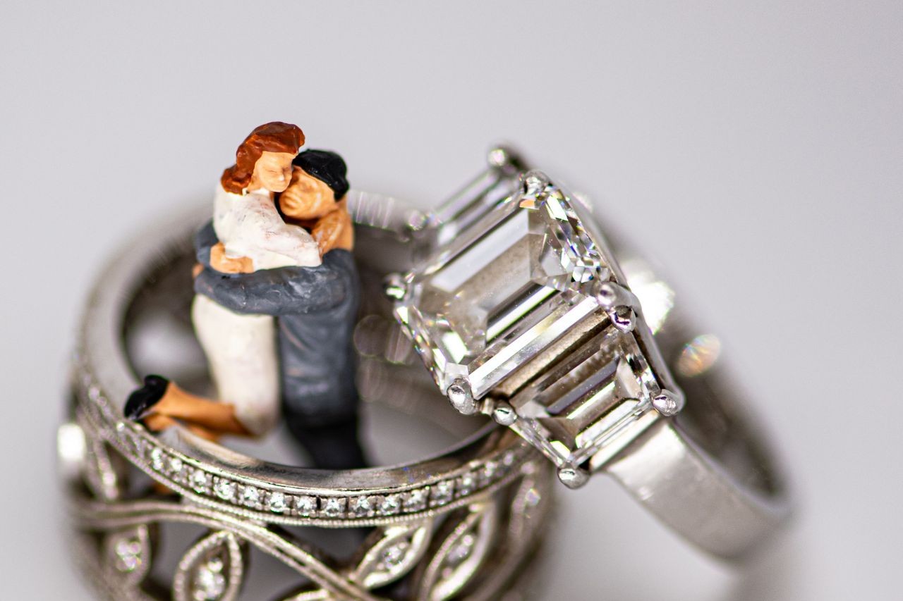 A close-up of two diamond rings, one featuring an intricate band design, with a miniature figurine couple embracing inside the ring.