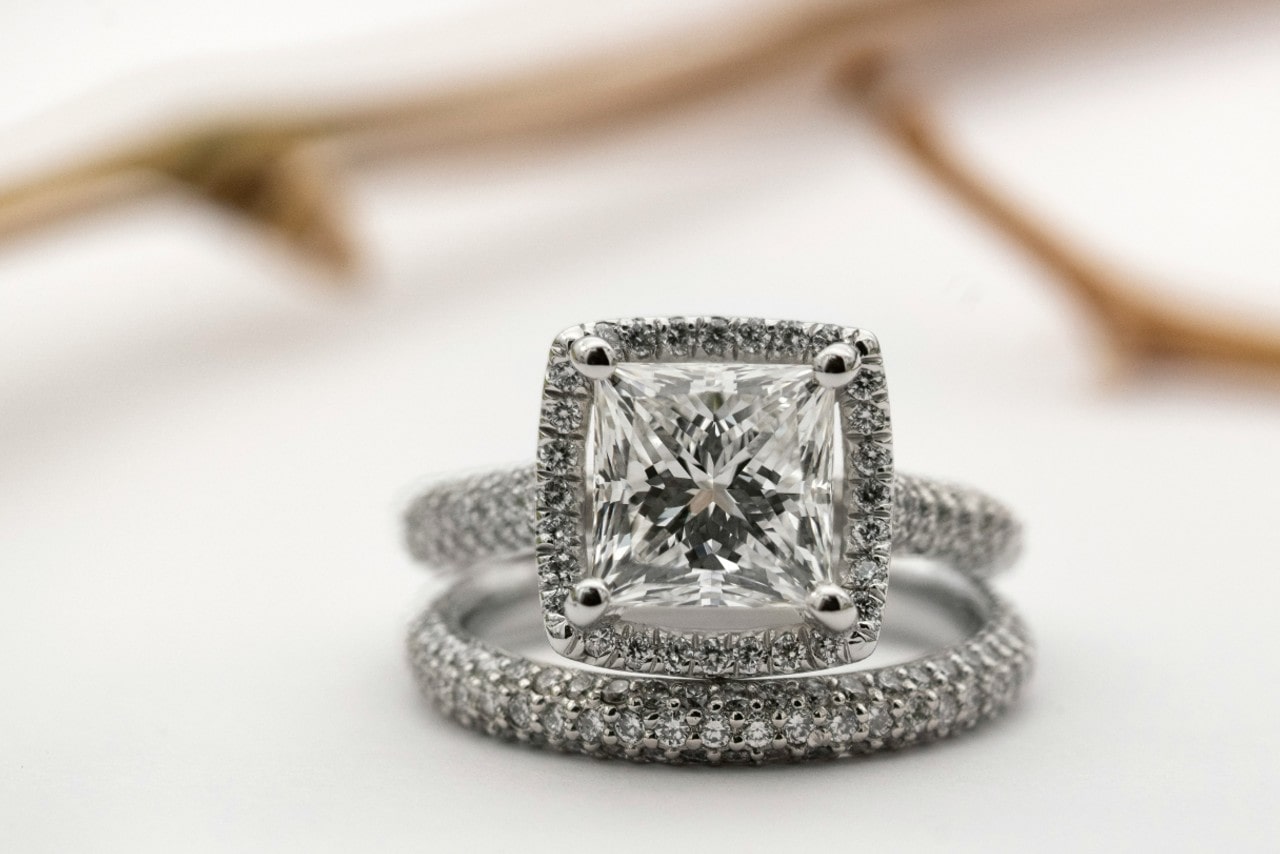 Close-up of an elegant cushion-cut diamond engagement ring with a pave-set band, paired with a matching wedding band, set on a soft white background.
