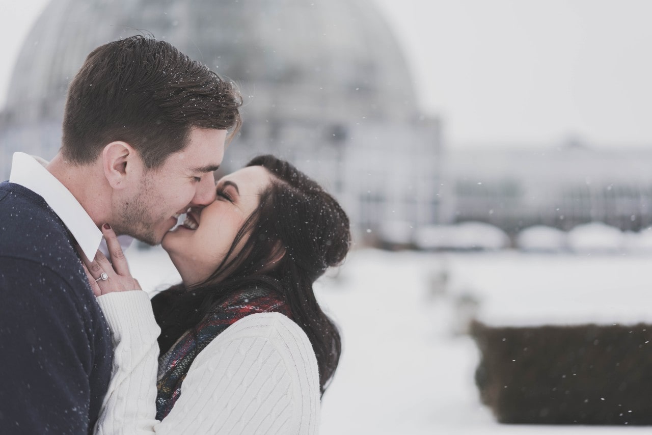 A couple embraces with smiling faces close, set against a snowy backdrop and blurred dome structure. A couple embraces with smiling faces close, set against a snowy backdrop and blurred dome structure.