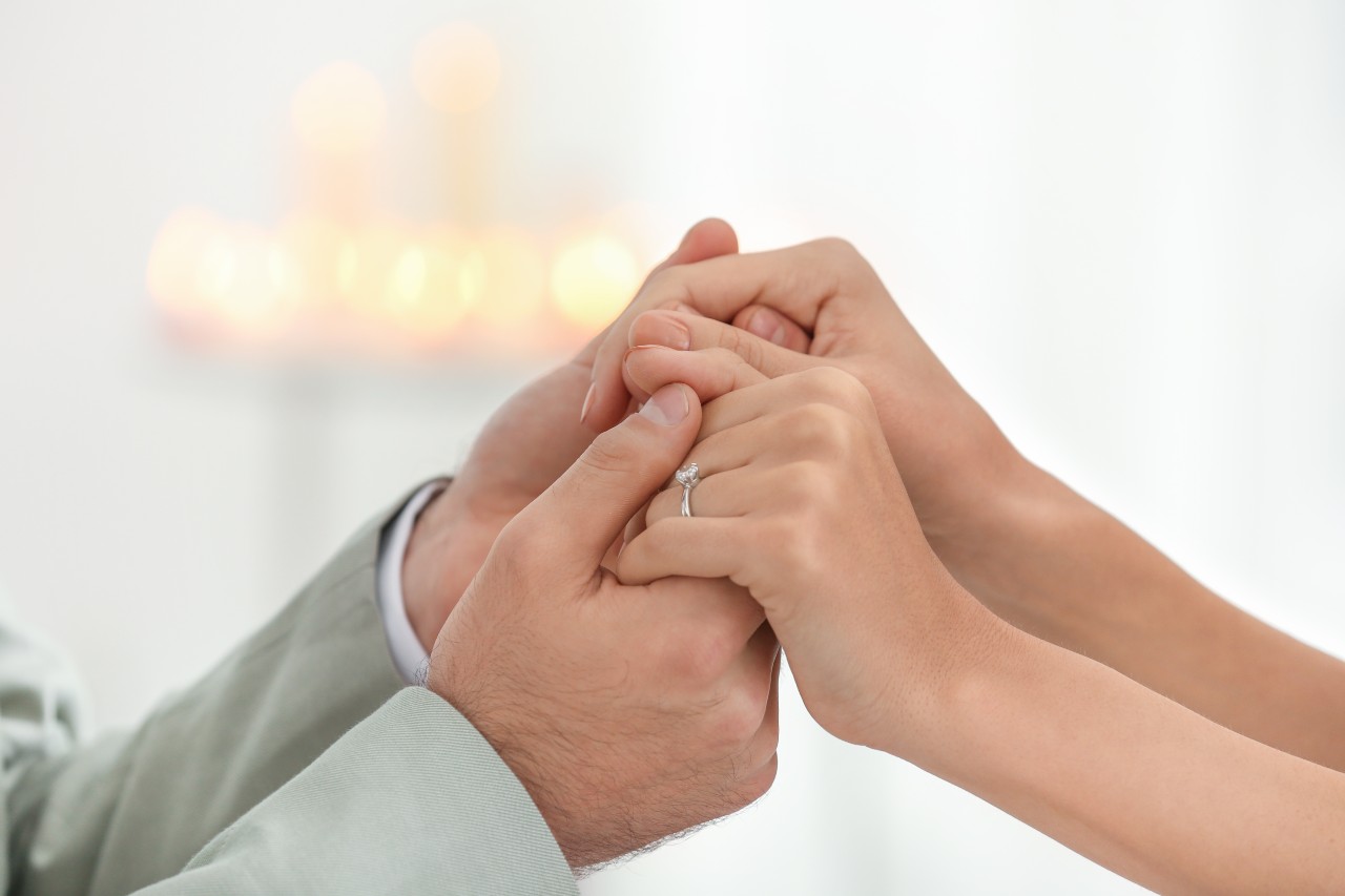 Close-up of two people holding hands, one wearing a solitaire engagement ring, with a soft, blurred background of glowing candles. Close-up of two people holding hands, one wearing a solitaire engagement ring, with a soft, blurred background of glowing candles.