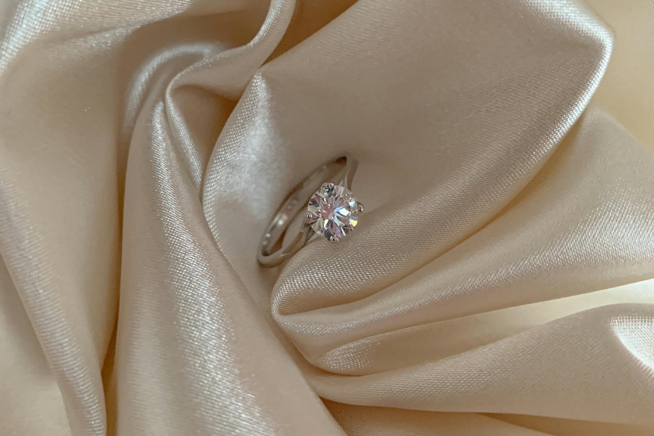 A sparkling solitaire diamond engagement ring rests on smooth, cream-colored satin fabric. A sparkling solitaire diamond engagement ring rests on smooth, cream-colored satin fabric.