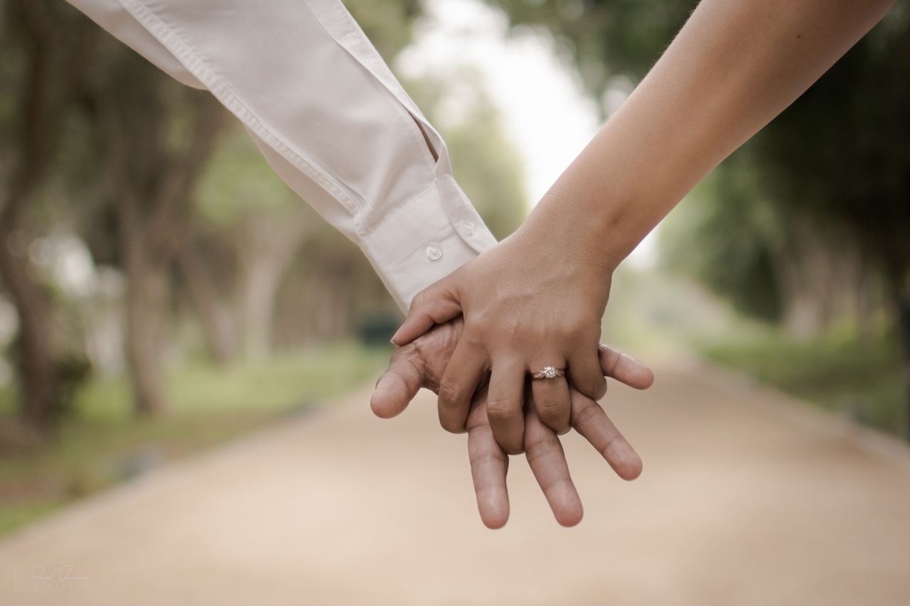 Two people hold hands on a blurred pathway, one wearing a white shirt and the other a yellow gold solitaire engagement ring. Two people hold hands on a blurred pathway, one wearing a white shirt and the other a yellow gold solitaire engagement ring.