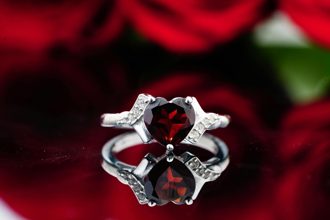 A silver ring with a heart-shaped deep red gemstone showcased against a blurry background of red roses A silver ring with a heart-shaped deep red gemstone showcased against a blurry background of red roses