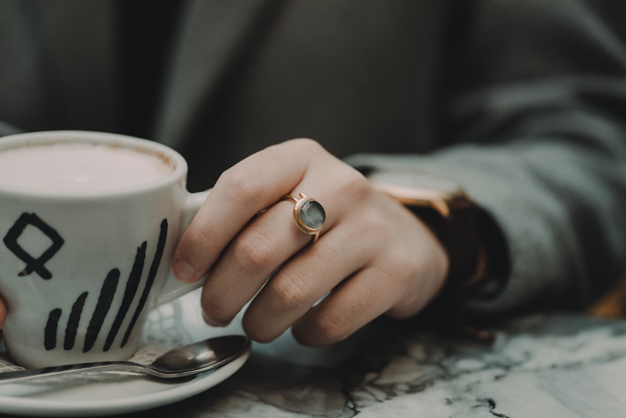 A close up of a hand with a gold ring with a round, translucent stone holding a ceramic mug of coffee A close up of a hand with a gold ring with a round, translucent stone holding a ceramic mug of coffee