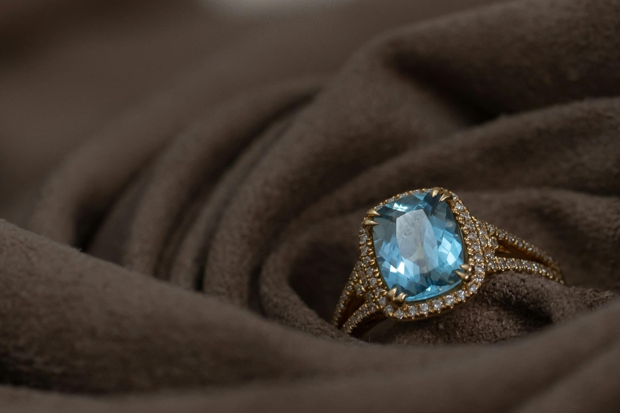 A gold ring with a large, sparkling blue gemstone surrounded by small diamonds, placed on soft brown fabric. A gold ring with a large, sparkling blue gemstone surrounded by small diamonds, placed on soft brown fabric.