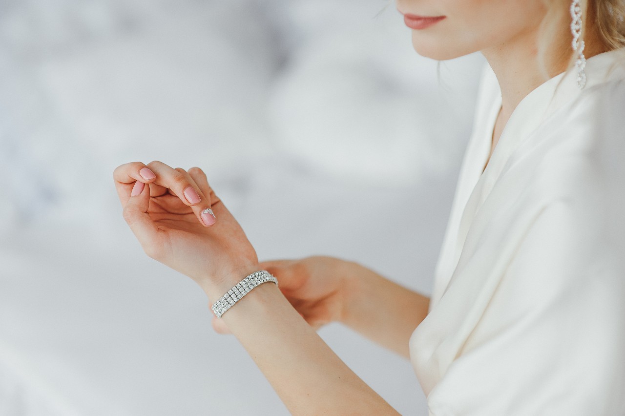 A woman wearing a white robe is putting on a diamond bracelet and is wearing dramatic drop diamond earrings