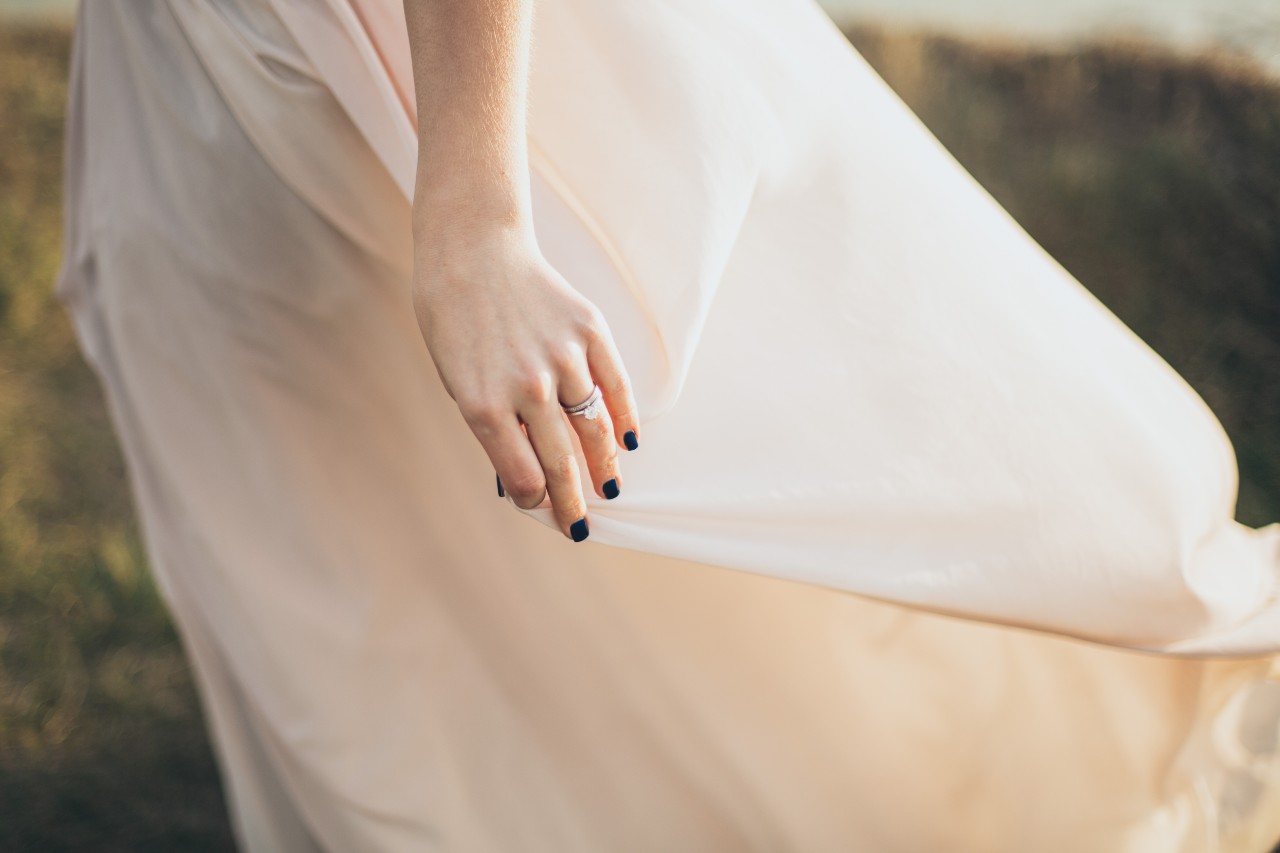 A bride standing outside, gently holding the skirt of her gown, wearing her engagement ring and wedding band together