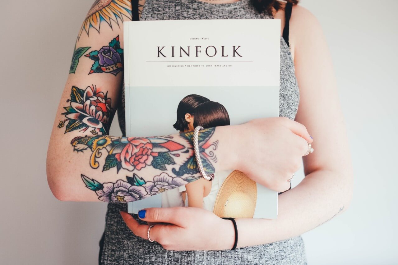 a tattooed woman wears a white gold bangle while holding a book titled “Kinfolk.”