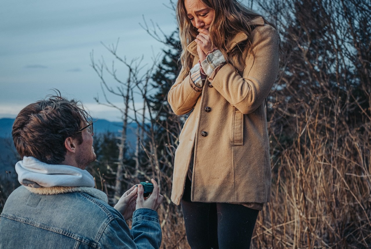 A man proposes to his girlfriend with a ring in an emerald-green box while they are taking an early morning hike in the wintertime
