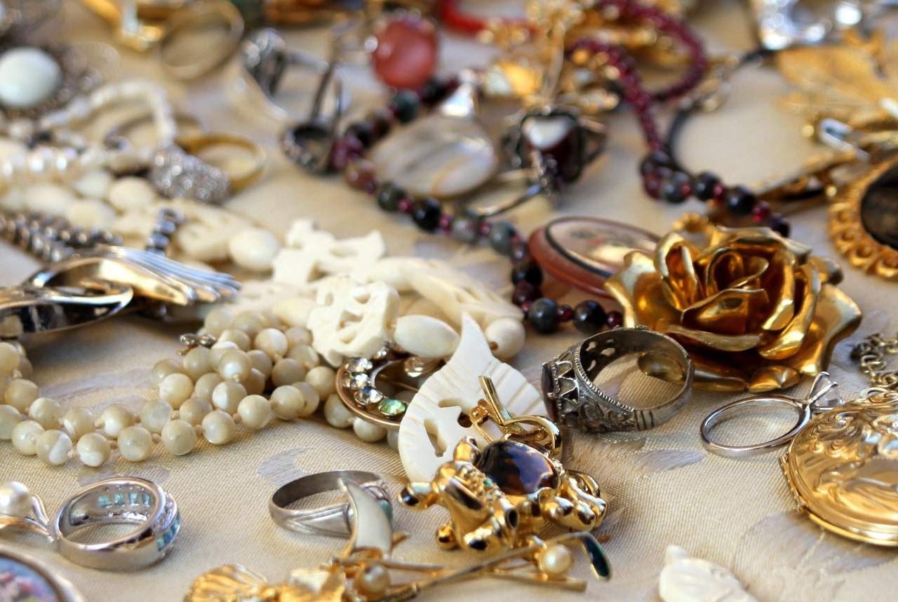 A huge collection of jewelry, featuring all types of styles, gemstones, and precious metals, are strewn across a tan tablecloth