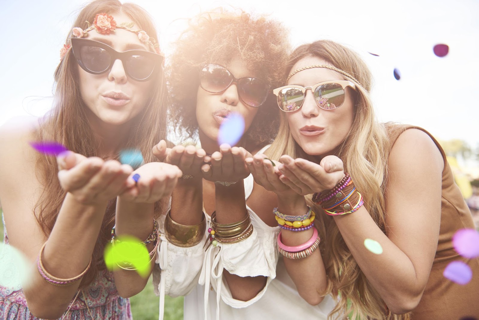Three women adorned with sunglasses, headbands, and bracelets blow confetti into the camera at a summer festival