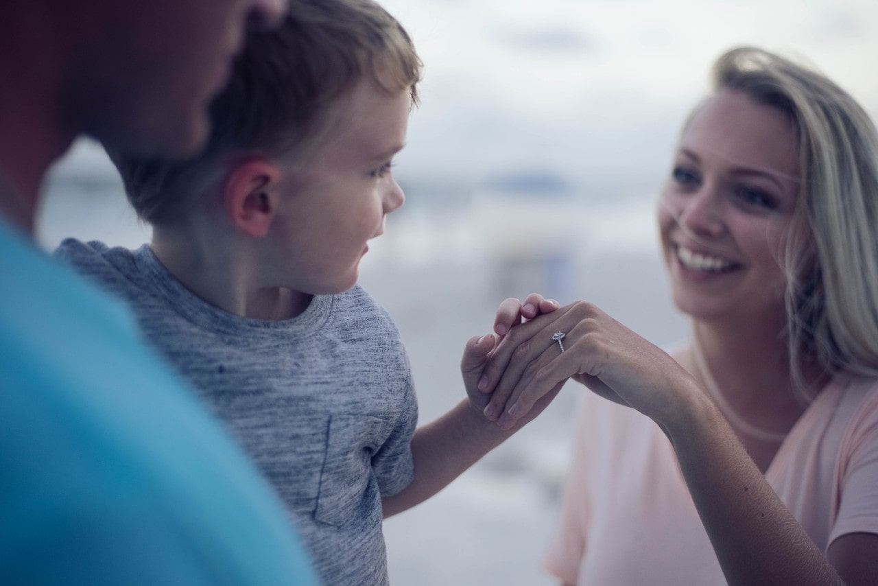 A mom shows her engagement ring to her young son on a beach