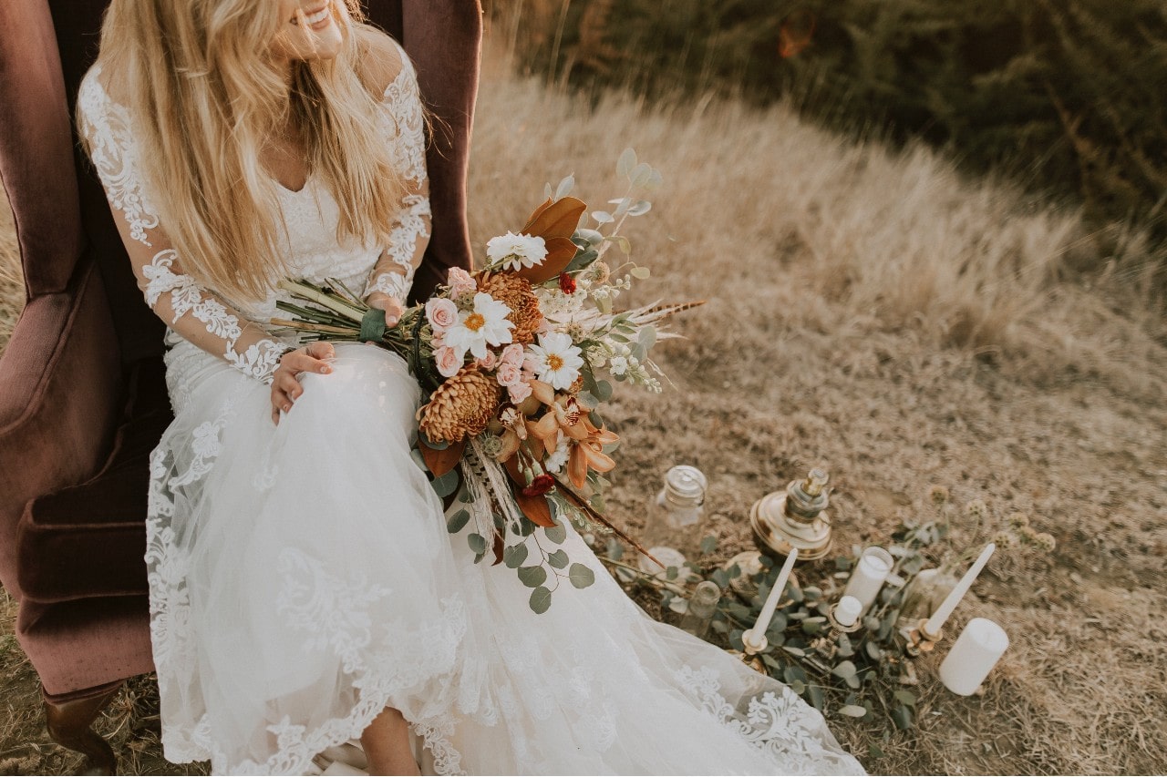 A bride sits on a chair with her bouquet in a field.