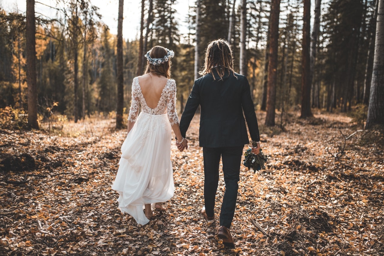 a bride and groom holding hands and walking through the woods in the fall
