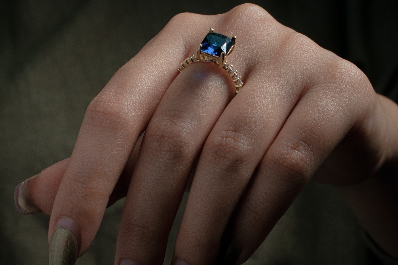 close up image of a woman’s hand wearing a gold fashion ring featuring a princess cut sapphire